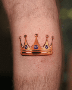 Gold crown tattoo with purple gemstones on a forearm, showcasing intricate design and vibrant colors, reflecting the artistic style of Pony Lawson at Inked Tattoo Studios.